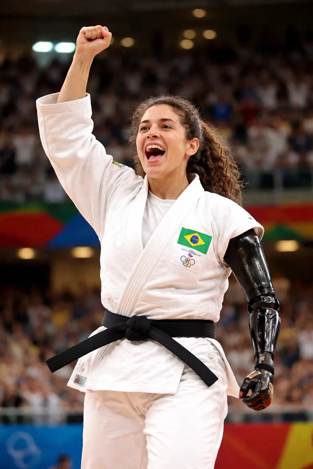 Uma atleta de judô com quimono branco e faixa preta, comemorando com o punho esquerdo erguido e a boca aberta em expressão de alegria. O braço direito é uma prótese mecânica preta, brilhante, com articulações visíveis. No peito do quimono há um patch com a bandeira do Brasil e, abaixo, pequenos símbolos dos anéis olímpicos; o texto é pequeno demais para leitura nítida. O cabelo é castanho e cacheado, preso para trás, e a pele é clara. Ao fundo há uma arena lotada e desfocada, com luzes fortes no alto.