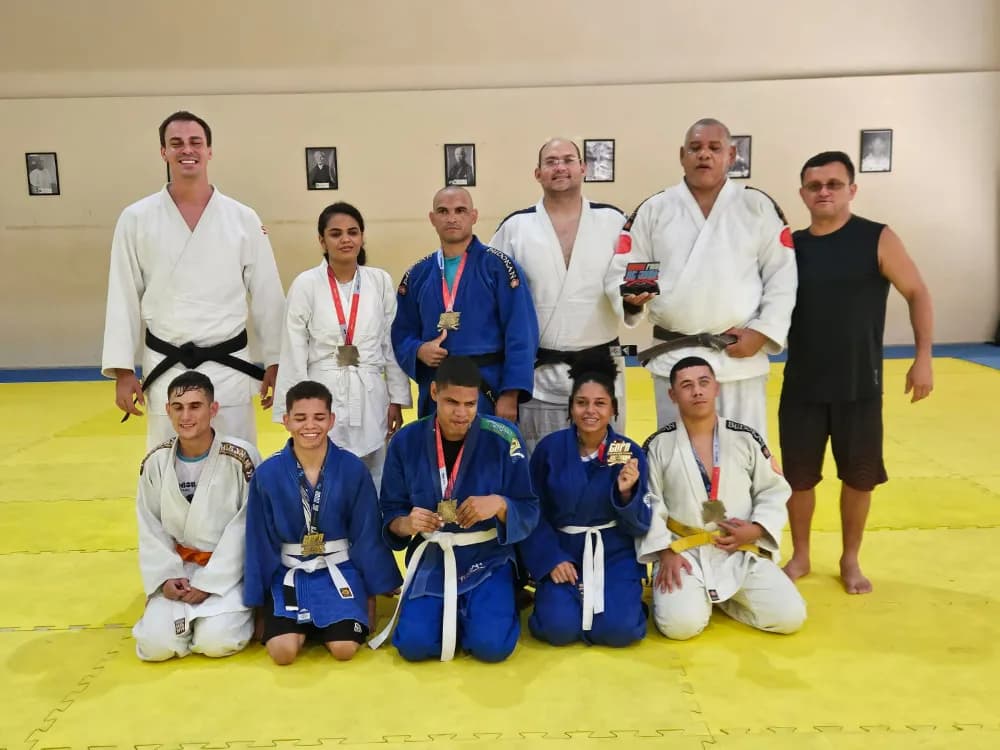 Um grupo de nove pessoas posando em um tatame amarelo, em um dojo, usando quimonos de judô (brancos e azuis) e exibindo medalhas.
Na fileira de trás há cinco pessoas em pé. Três homens de quimono branco (um bem alto com faixa preta; outro de estatura média com faixa preta; e um homem mais alto e mais largo com faixa preta segurando um troféu/placa). Entre eles há um homem de quimono azul com medalhas no pescoço. Ao lado esquerdo, há uma mulher de quimono branco com medalha. À extrema direita, há um homem sem quimono, usando camiseta preta sem mangas e bermuda escura.
Na fileira da frente há quatro pessoas ajoelhadas. Um homem de quimono branco à esquerda, com medalha e faixa laranja. Três pessoas de quimono azul e uma de quimono branco à direita (com faixa amarela), todas com medalhas; uma das pessoas na frente segura uma medalha voltada para a câmera.