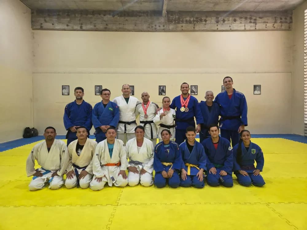 Grupo de judocas posando em tatame amarelo e azul.
Há 16 pessoas ao todo, organizadas em duas fileiras: oito agachadas na frente e oito em pé atrás.
Todas estão usando quimono de judô, em cores branca ou azul, com faixas de diferentes cores na cintura, indicando graduações variadas (branca, amarela, laranja, azul, roxa e marrom).
No centro da fileira de trás, um homem de quimono branco e faixa preta está ligeiramente à frente dos outros, passando a impressão de ser o instrutor ou professor.
À direita dele (para quem observa), um homem de quimono azul exibe várias medalhas douradas e vermelhas penduradas no pescoço e sorri.
Ao lado desse homem com medalhas há uma mulher de cabelo grisalho curto, também sorridente, de quimono azul escuro.
No meio do grupo da frente há uma mulher de quimono branco, sentada de joelhos, também sorridente.
As expressões do grupo em geral são de satisfação e orgulho, como se tivessem participado ou acabado de sair de uma competição ou exame de graduação.
O ambiente é um dojô interno, com paredes claras e teto de concreto aparente, e nas paredes ao fundo há pequenos quadros ou fotos emolduradas alinhadas horizontalmente.